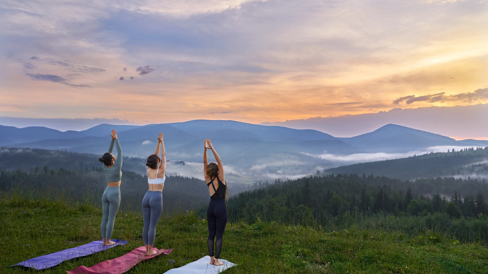 Tre donne praticano yoga sui tappetini su un prato, davanti alle montagne all'alba